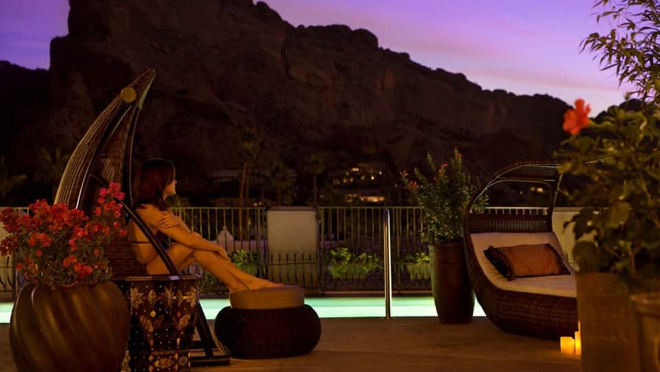 Woman relaxing in lounge chair on terrace at dusk with rock formations and illuminated pool behind