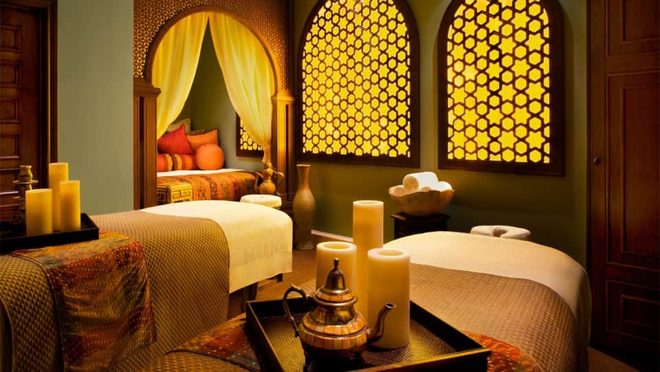 Moroccan-style spa treatment room with two massage beds, arched windows, candles, and golden lighting
