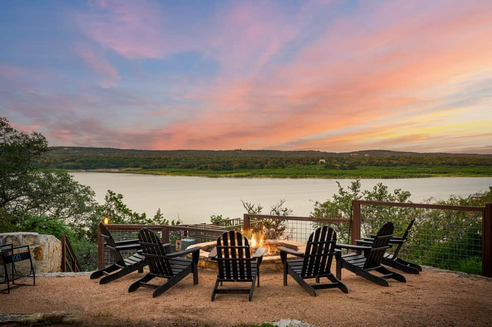 Sunset over a tranquil lake with Adirondack chairs arranged around an outdoor fire pit