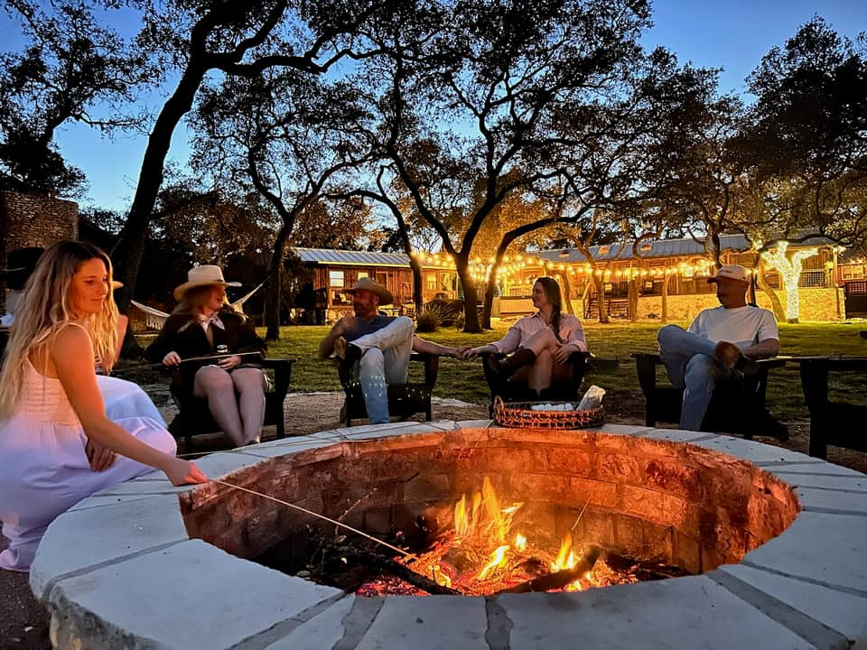 Evening gathering around a large fire pit with oak trees and illuminated ranch buildings