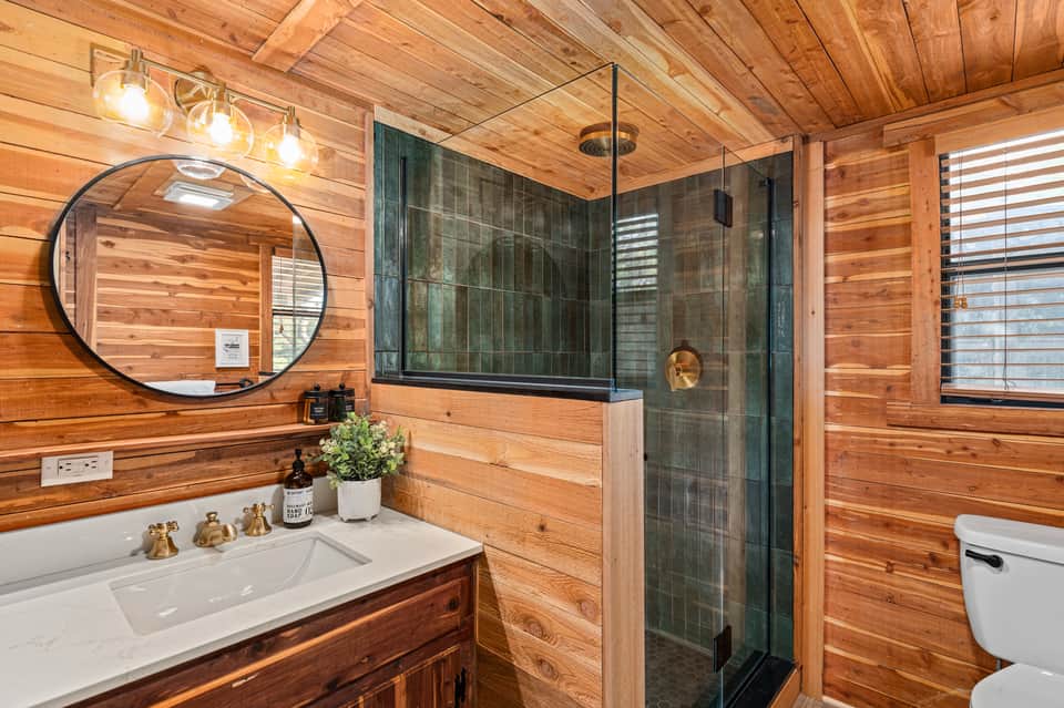 Modern wooden bathroom with stone shower, white vanity sink, and circular mirror lighting