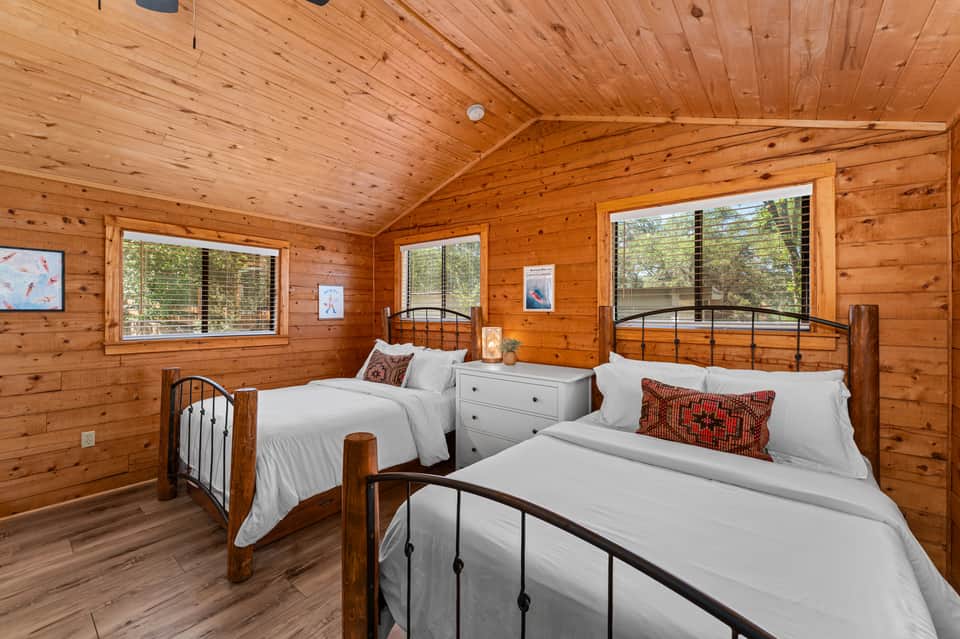 Spacious wooden cabin bedroom with two metal-frame beds, white linens, red pillows, and large windows overlooking trees