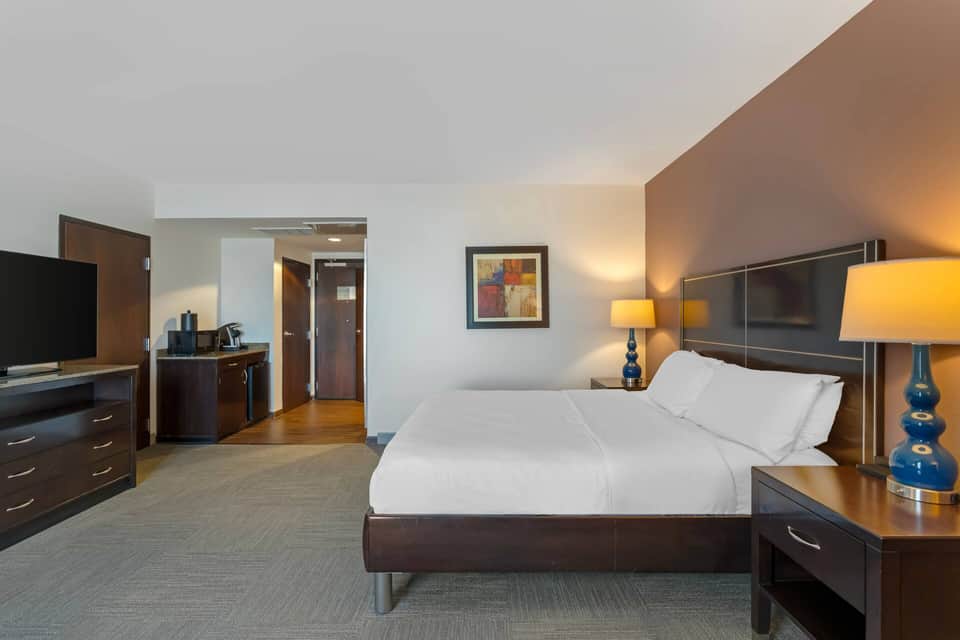 Spacious hotel room with dark wood bed frame, flat-screen TV, desk area, and warm amber lighting