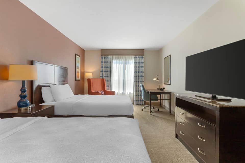 Bright double queen room with mauve accent wall, work desk, flat-screen TV, and large windows with curtains