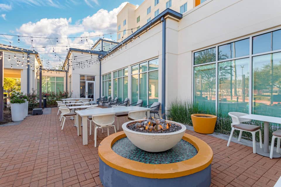 Upscale outdoor patio with fire pit, dining tables, string lighting, and glass doors to hotel interior