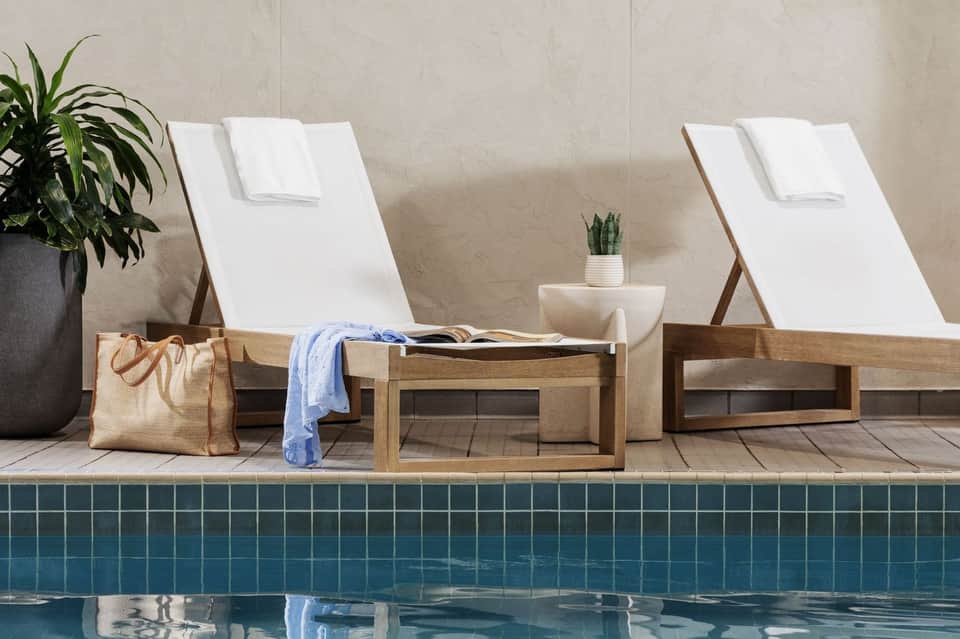 Two wooden loungers with white cushions beside an indoor pool with potted plants