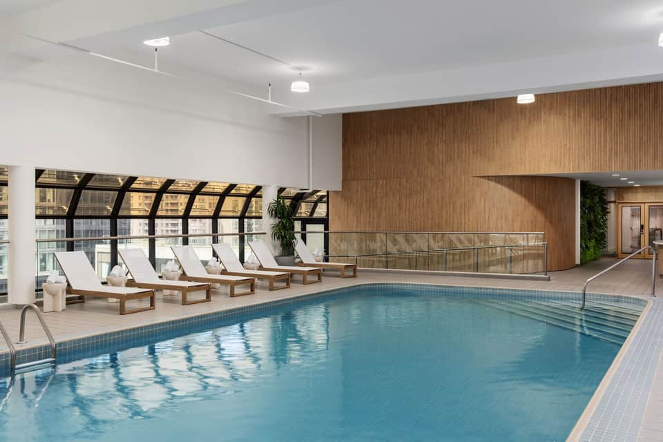 Indoor pool with wooden loungers, white cushions, and floor-to-ceiling windows in luxury facility