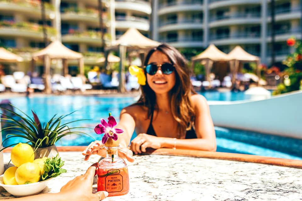 Woman in sunglasses at resort pool holding Patrón tequila bottle with lemons and flowers