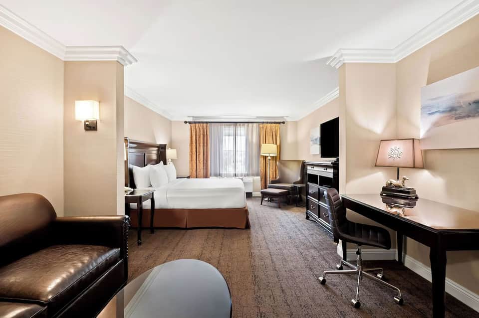 Spacious hotel suite with king bed, seating area, fireplace, dark wood furniture, and work desk