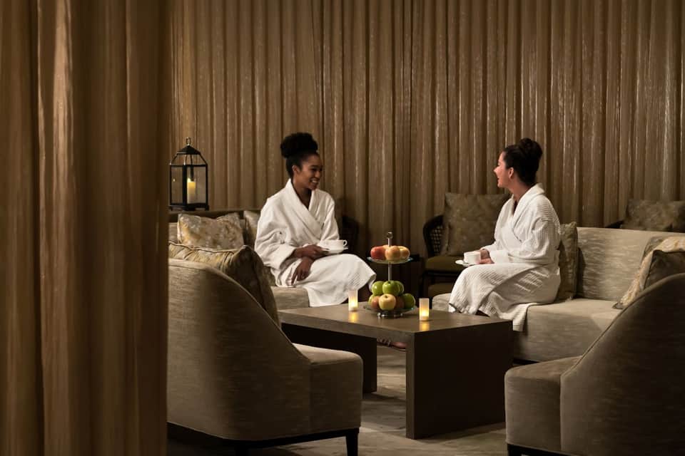 Two women in white robes sitting on chairs having conversation in spa lounge area