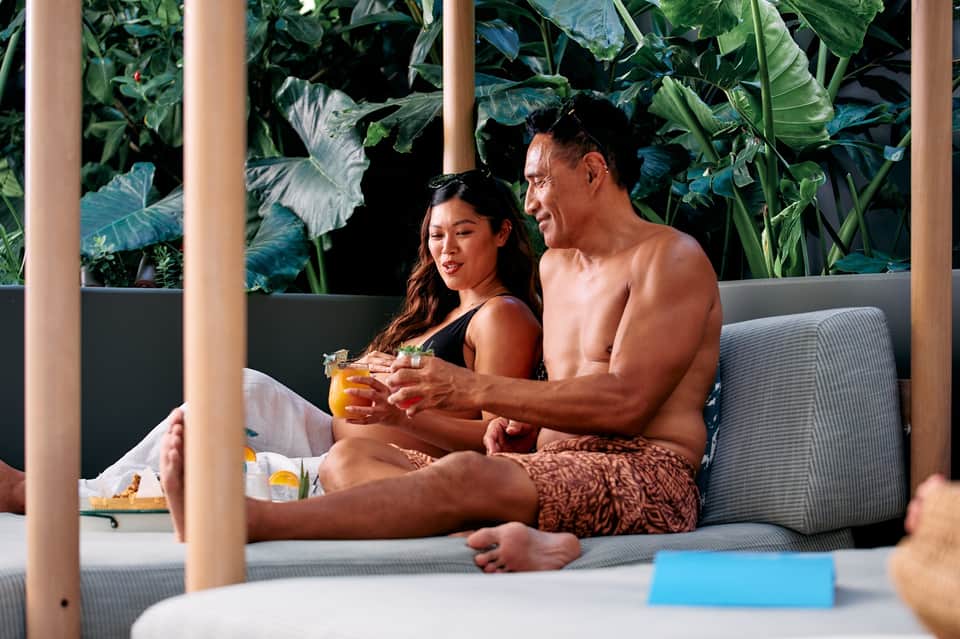 Couple relaxing on a lounge chair with tropical foliage backdrop, holding cocktails at resort