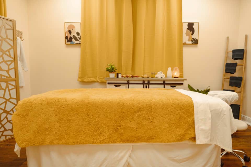 Spa room with golden yellow curtains, massage bed with mustard blanket, shelving, and artwork