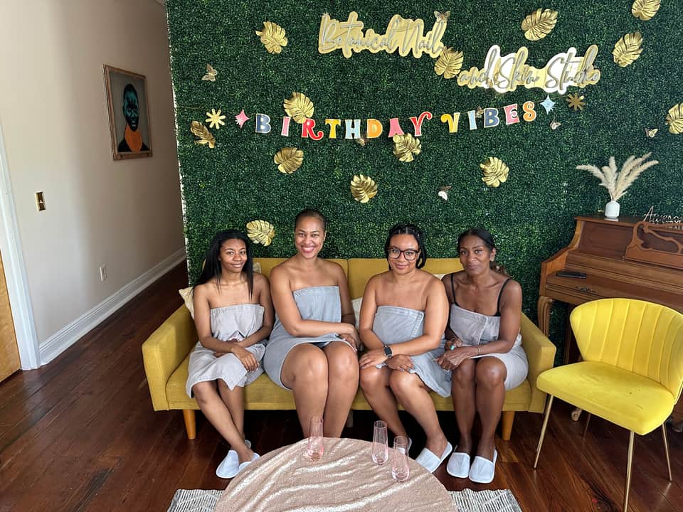 Four women in spa robes sitting on a yellow couch in front of a green botanical backdrop with birthday signage