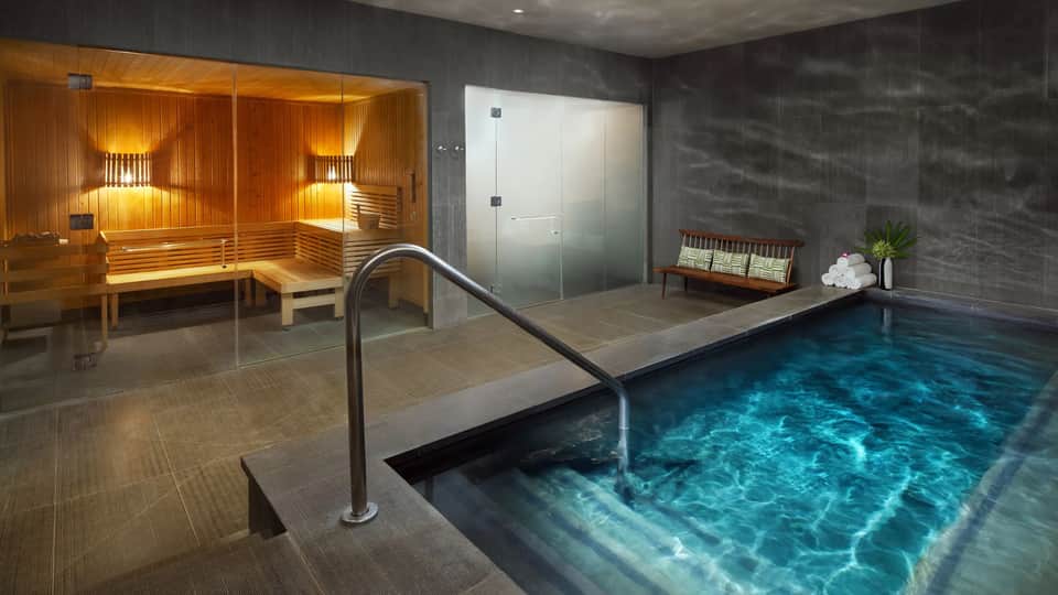 Indoor spa area with illuminated sauna, frosted glass shower enclosure, and turquoise swimming pool with metal handrail