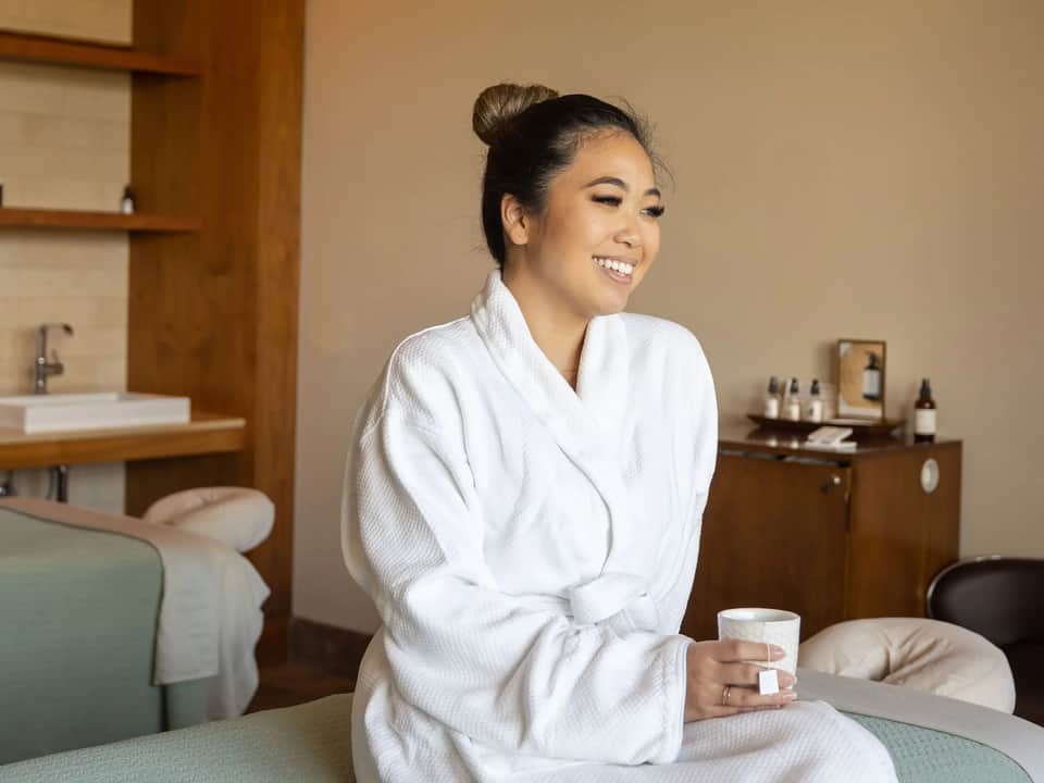 Woman in white robe smiling in spa treatment room with wooden accents, sink, and skincare products on shelf