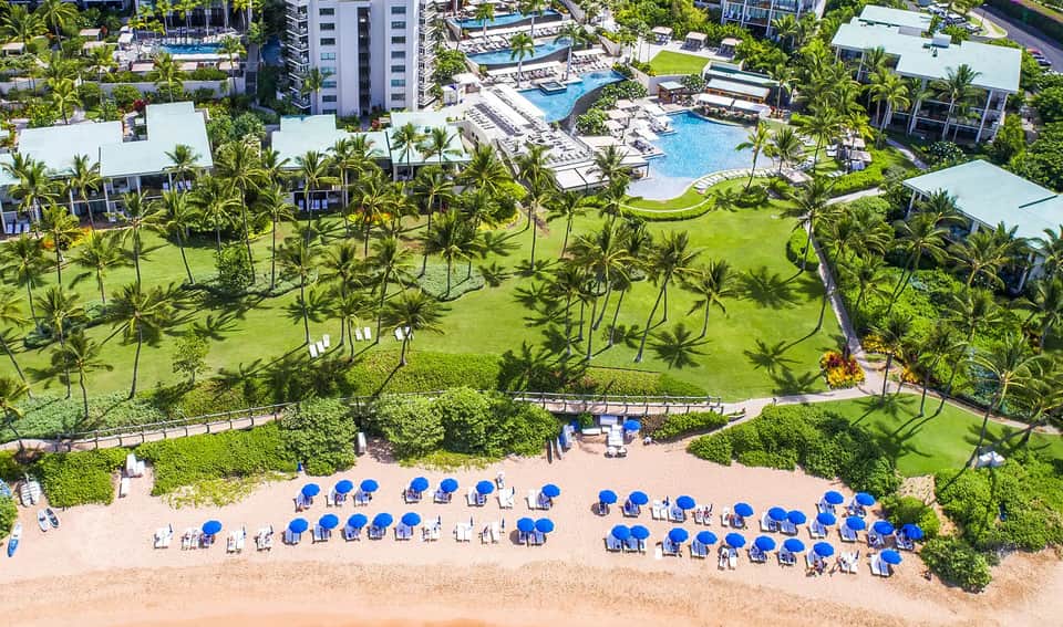 Aerial view of beachfront resort with pools, manicured gardens, palm trees, and rows of blue umbrellas on sandy beach