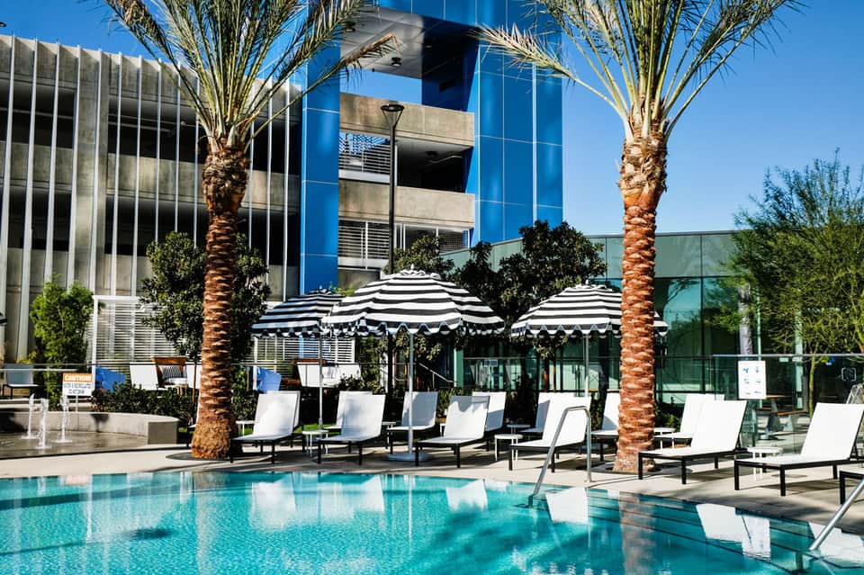 Resort pool deck with striped umbrellas, lounge chairs, and palm trees under clear blue sky