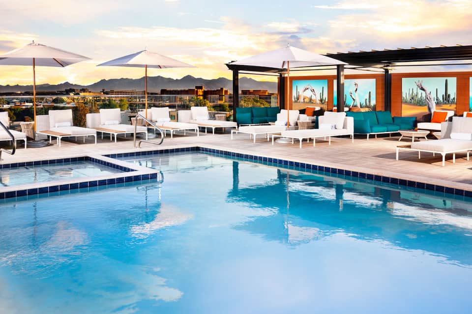 Resort pool deck at sunset with lounge chairs, umbrellas, and colorful modern buildings in background