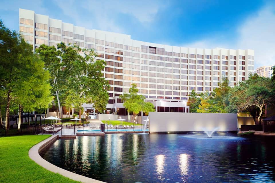 Modern high-rise hotel building beside illuminated fountain and curved pond with manicured lawn and trees