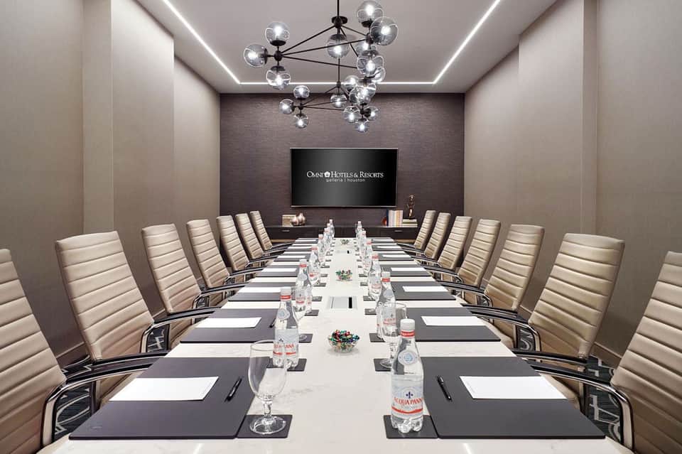 Modern conference room with long table, beige chairs, chandelier, and branded screen display