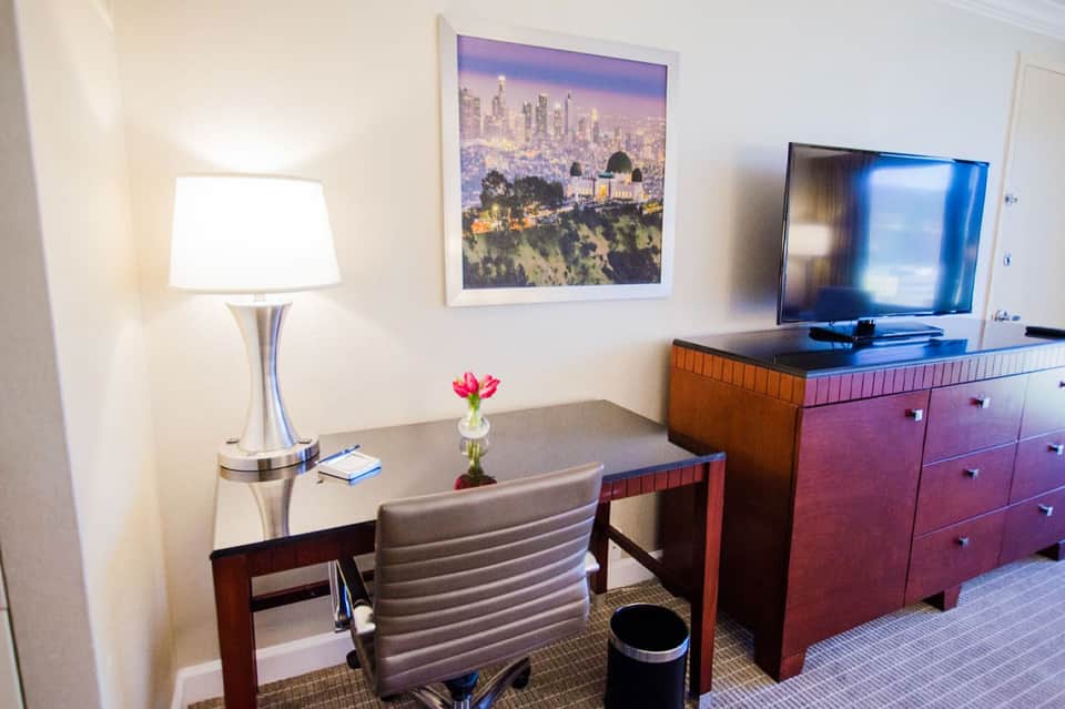 Modern hotel room with desk, gray chair, flat-screen TV, wood dresser, and cityscape artwork on wall