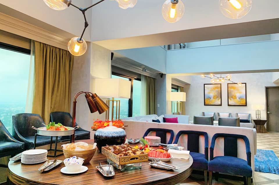 Modern hotel suite with pendant lights, lounge seating, round coffee table with snacks, and city views