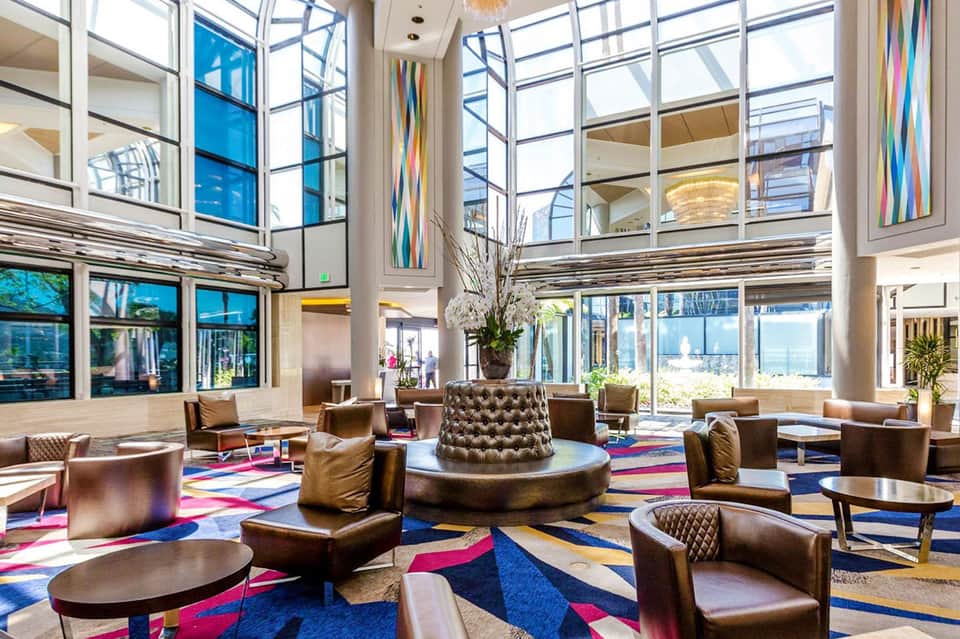Modern hotel lobby with high ceilings, floor-to-ceiling windows, colorful carpet, leather seating, and natural light