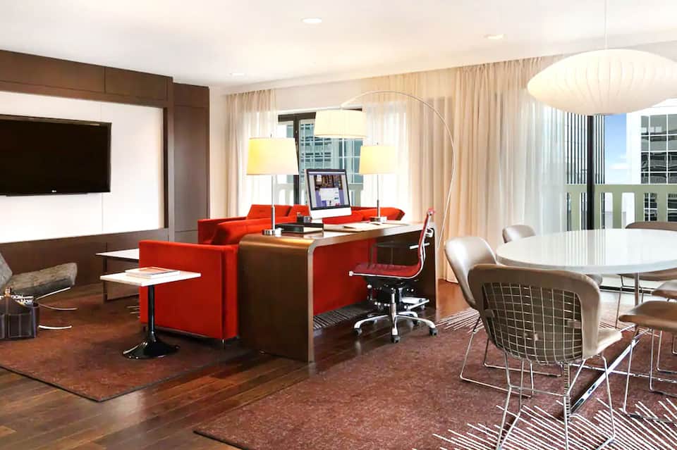 Modern hotel suite with red accent chairs, wooden desk, and floor-to-ceiling windows