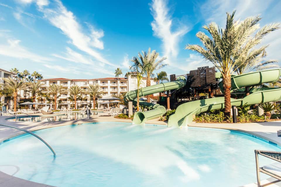 Resort pool with water slides, palm trees, and multi-story hotel building under blue sky