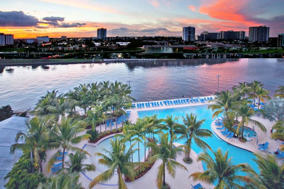 Resort pool complex at sunset with palm trees, multiple pools, and waterfront cityscape