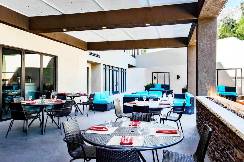 Modern courtyard dining area with round tables, blue cushioned seating, and covered pergola with fireplace
