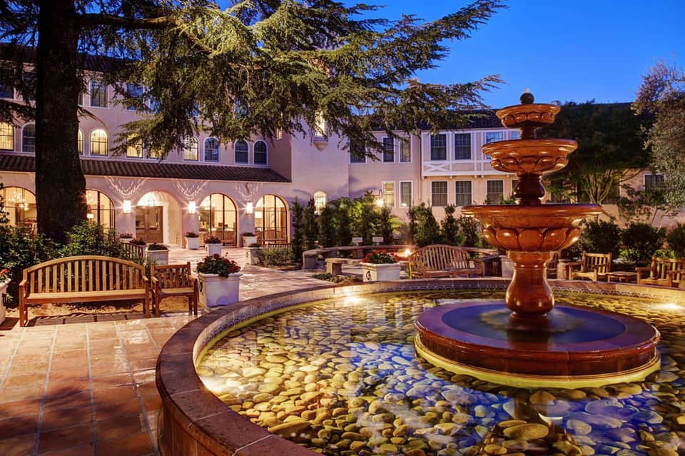 Elegant courtyard with illuminated fountain, arched colonnade, wooden benches, and pink stucco buildings at dusk