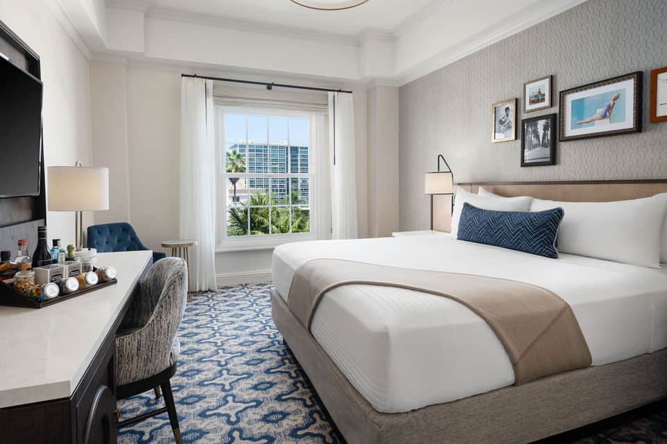 Upscale hotel bedroom with king bed, desk, and floor-to-ceiling windows overlooking city skyline