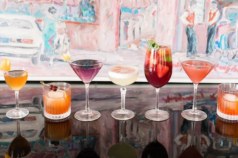 Colorful cocktails displayed on a reflective bar counter with abstract artwork backdrop