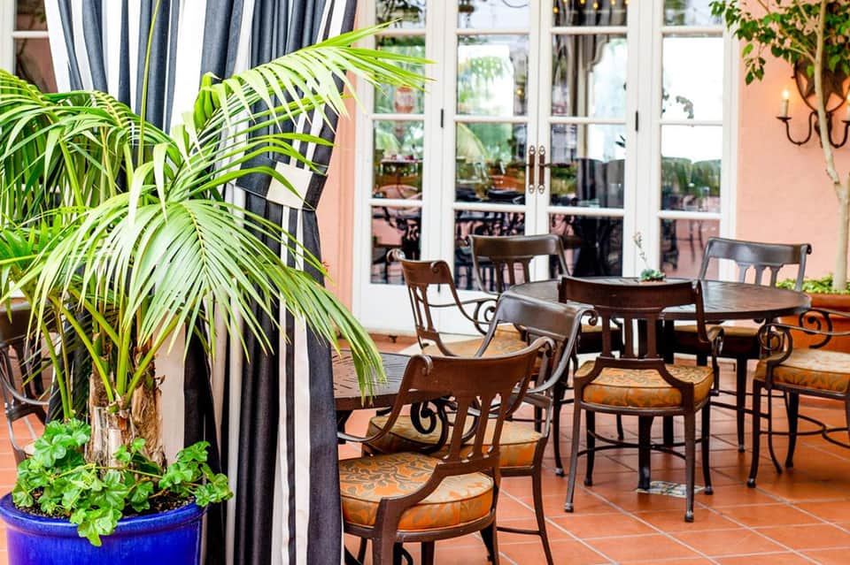 Elegant indoor lounge with palm plants, striped curtains, wooden chairs, and glass doors opening to terrace