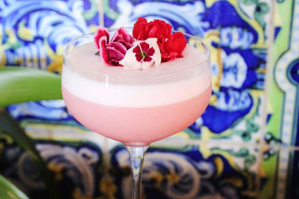 Pink cocktail with layered cream and red flower garnish against decorative blue and yellow tile backdrop