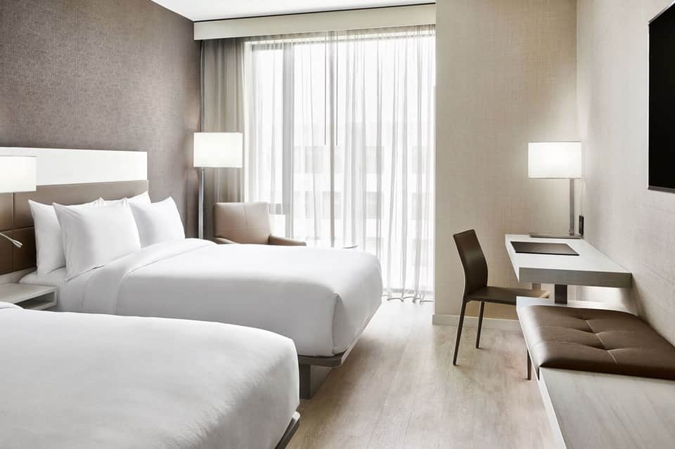Modern hotel room with two queen beds, work desk, and floor-to-ceiling windows with sheer curtains