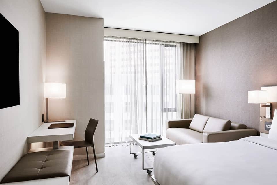 Modern hotel room with neutral tones, queen bed, work desk, sofa, and floor-to-ceiling windows with sheer curtains