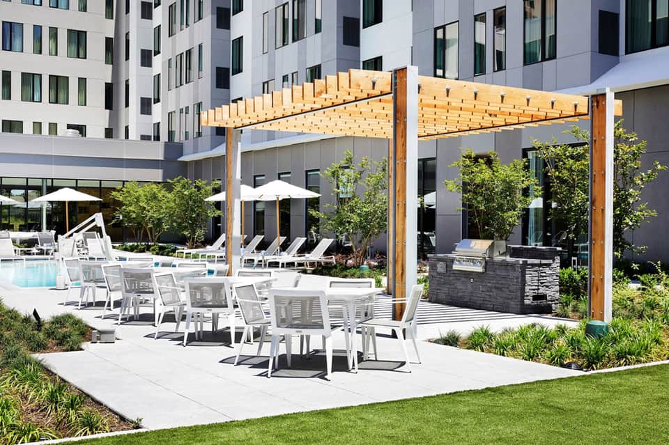 Modern resort courtyard with wooden pergola, dining tables, lounge chairs, pool, and high-rise buildings