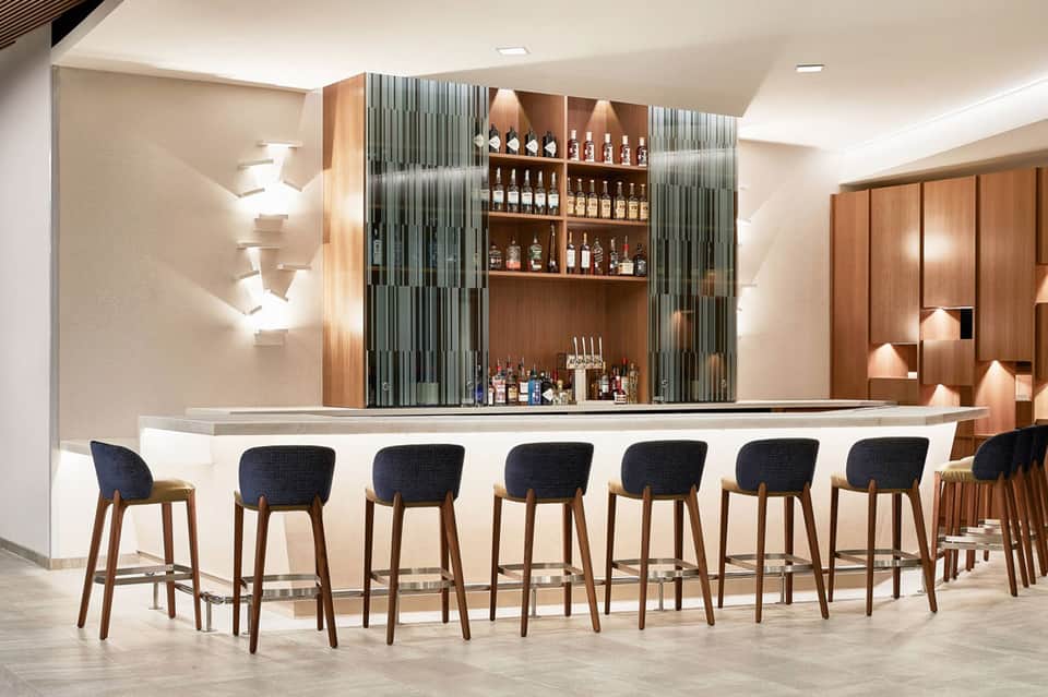 Modern hotel bar with curved white counter, wooden back shelving with bottles, and dark upholstered bar stools with wooden legs