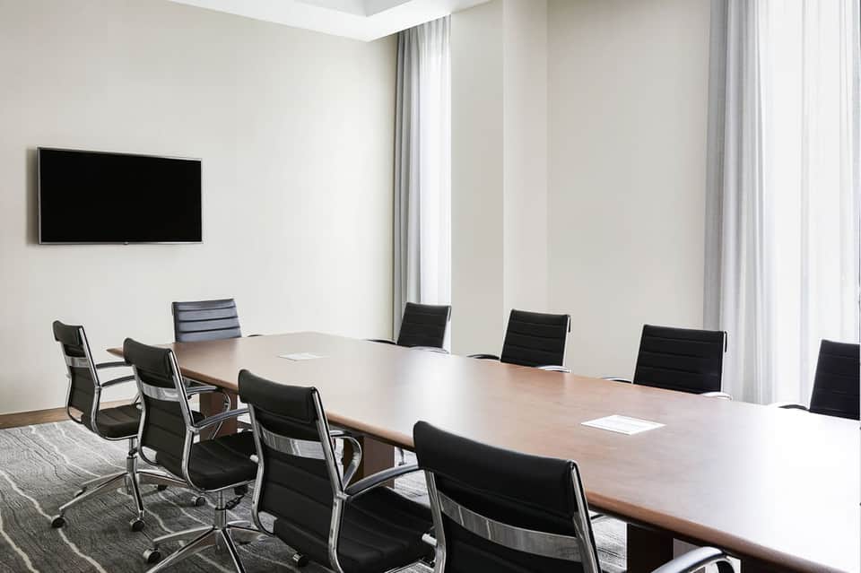 Contemporary conference room with long wooden table, black office chairs, wall-mounted TV, and floor-to-ceiling windows