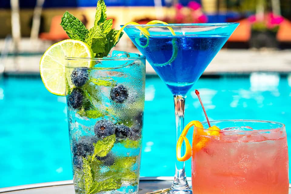 Three colorful cocktails by a turquoise pool with fresh berries, lime, and mint garnish