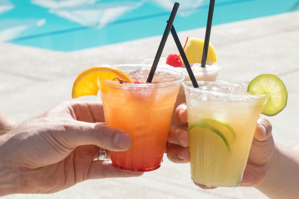 Two colorful tropical cocktails by a poolside with citrus garnishes and black straws