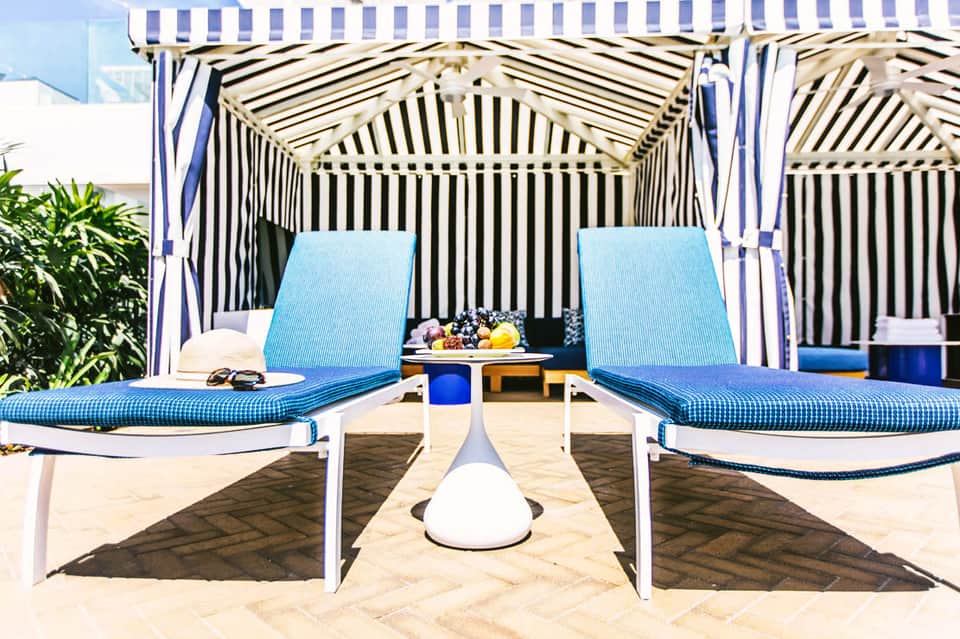 Two blue lounge chairs under striped cabanas on a sandy beach with white and blue striped umbrellas