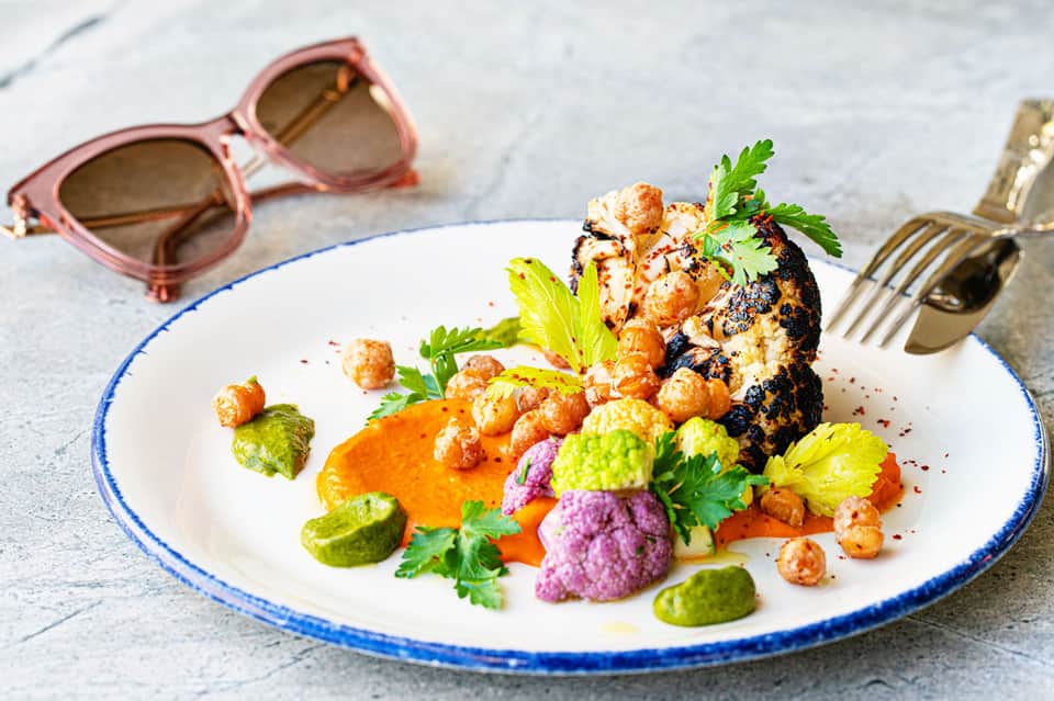 Artfully plated dish with seared fish, colorful roasted vegetables, and edible flowers on white plate with pink sunglasses nearby
