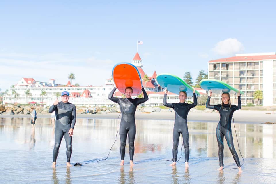 Surfers in wetsuits holding colorful surfboards on a beach near a Victorian-style building