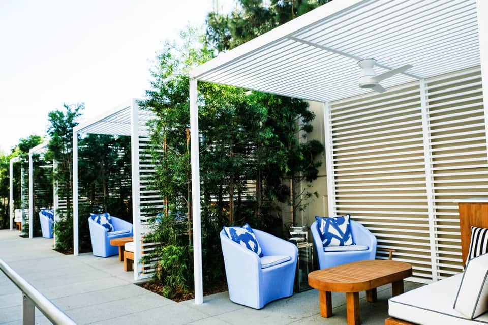 Outdoor patio seating area with blue accent chairs, wooden tables, white pergola, and green planted privacy screens