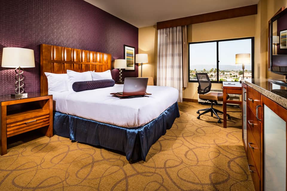 Upscale hotel bedroom with wooden headboard, purple accent wall, white bedding, work desk, and city view through large windows