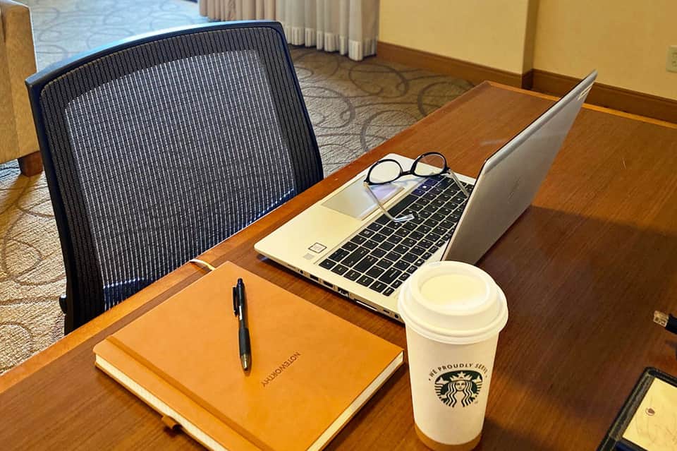 Hotel workspace with laptop, Starbucks coffee, orange notebook, and glasses on wooden desk