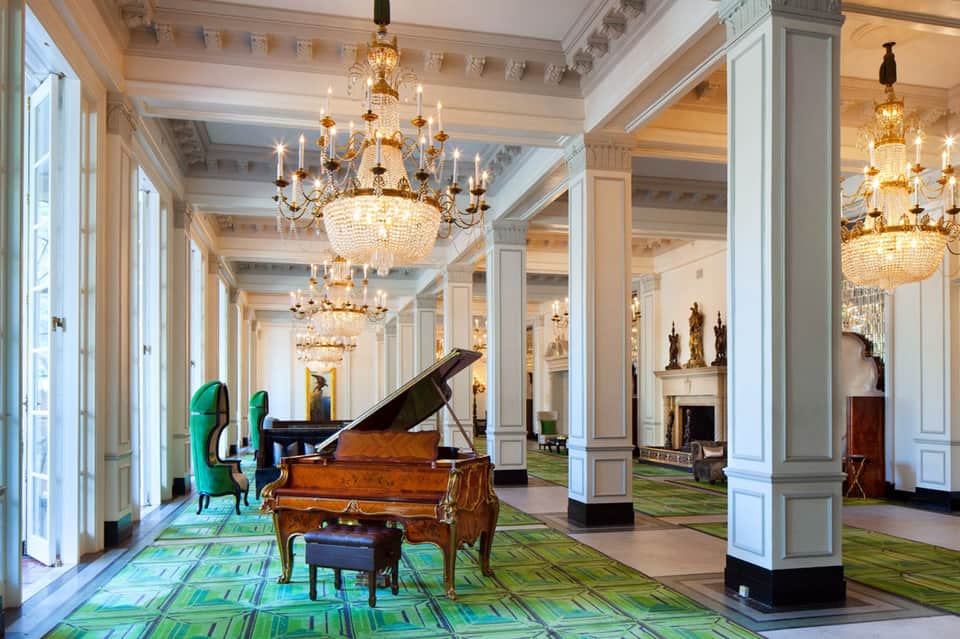 Elegant hallway with crystal chandeliers, grand piano, white columns, and green patterned carpet in luxury hotel